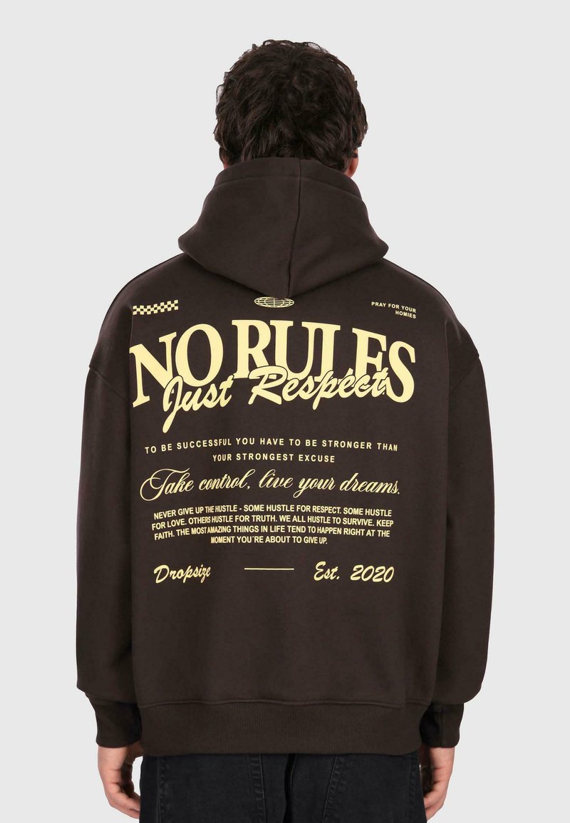 Personne portant un sweat à capuche noir avec du texte jaune au dos : "PAS DE RÈGLES Juste du Respect," phrases motivantes, et "Dropsiz Est. 2020."