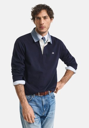 Polo à manches longues bleu marine avec un col et des poignets bleus clairs en contraste, arborant un petit logo brodé, associé à un jean bleu clair.