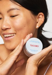 Crème Multi-Céramide dans un pot rond blanc avec des inscriptions bleues et rouges. Texture lisse ; 50 ml. Le modèle tient le pot, montrant une peau propre et un léger sourire.