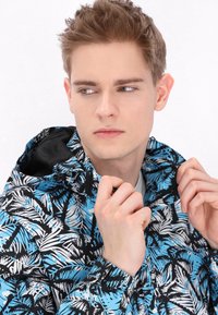 Jachetă cu glugă neagră și albastră, cu un model de frunze tropicale; material ușor, închidere cu fermoar și glugă ajustabilă, arătată parțial cu fermoarul închis.