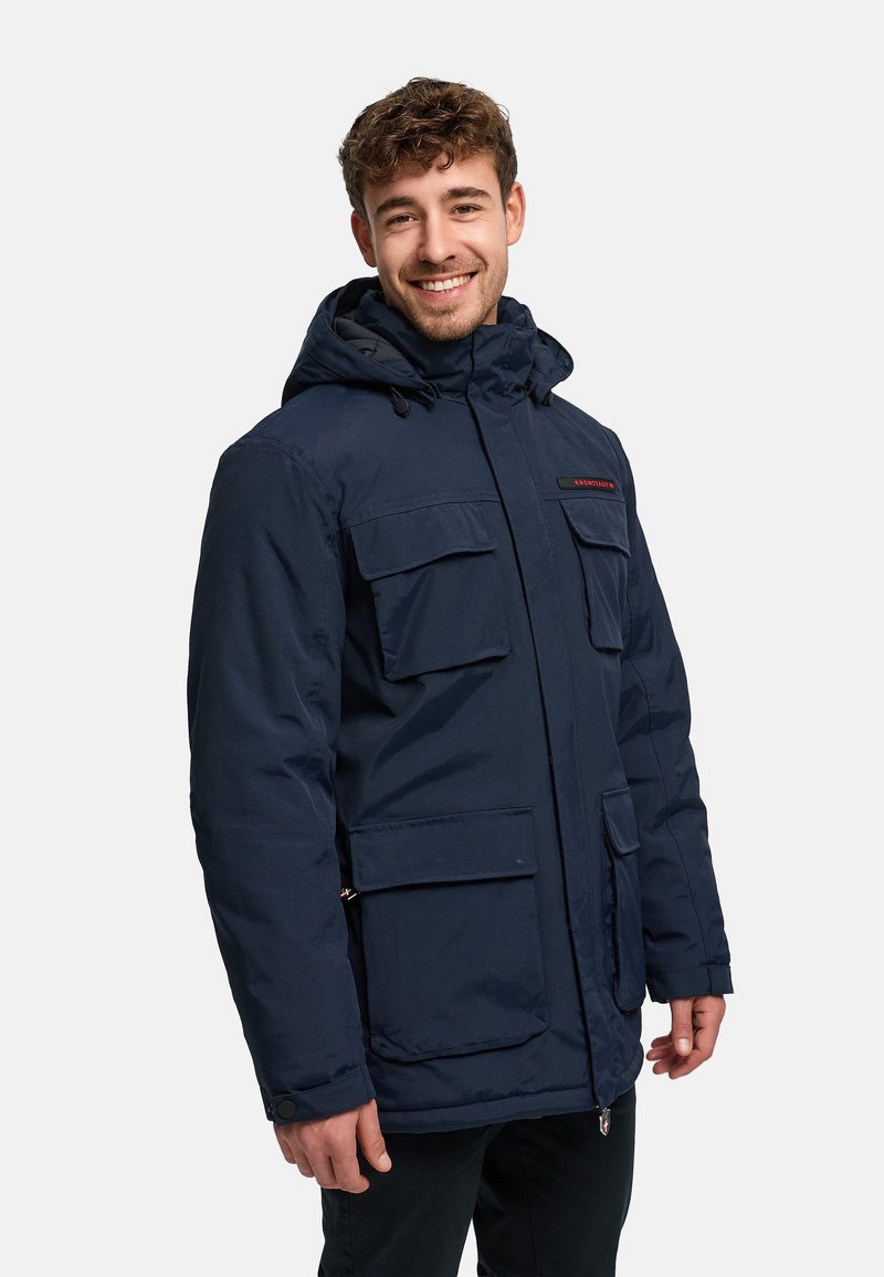 Parka d'hiver marine avec capuche, deux grandes poches avant et une fermeture éclair. Présente un écusson logo sur la poitrine.