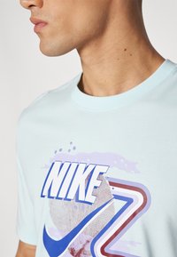 Bavlněné tričko světle modré barvy s vícebarevným grafickým prvkem, obsahujícím text "NIKE", zakřivené akcenty a texturovaný vzor v designu.