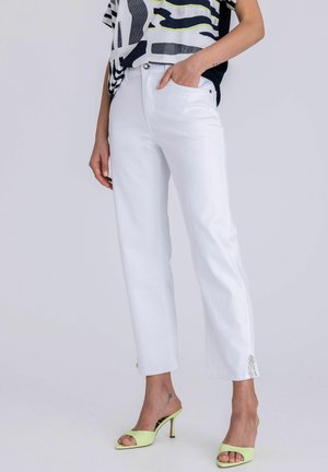 Persoon draagt witte rechte jeans met kleine kettinkjes aan de zoom, lichtgroene open teen sandaaltjes met hak en een zwart-wit patroon top.