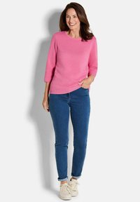 Eine lächelnde Frau trägt einen strukturierten rosa Pullover, blaue Jeans mit hochgekrempelten Säumen und weiße Sneakers, und steht vor einem schlichten hellen Hintergrund.