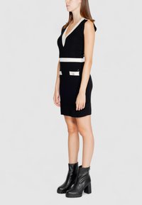 Schwarzes Kleid mit tiefem V-Ausschnitt, weißem horizontalem Band in der Taille und zwei Vordertaschen. Getragen mit schwarzen klobigen Absatzstiefeln.