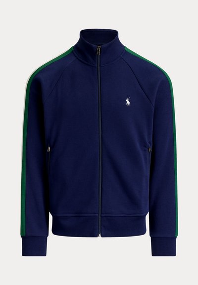 Veste de survêtement bleu marine avec fermeture éclair, bandes vertes sur les manches et logo joueur de polo blanc sur la poitrine gauche.