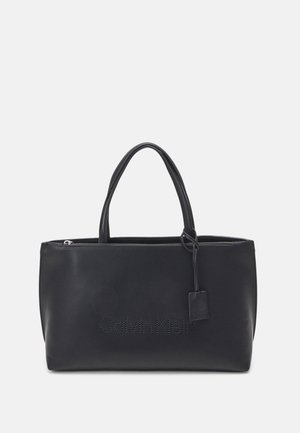 Czarna prostokątna torba typu tote z dwoma uchwytami, zamkiem błyskawicznym i perforowanym logo "Calvin Klein" na przodzie.