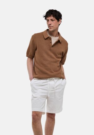 Homme portant un polo marron à manches courtes et un short blanc à cordon, debout les mains dans les poches devant un fond uni.