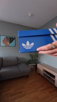 Caixa de sapatos Adidas em cartão azul com logótipo branco e design de três riscas. A caixa é segurada à mão contra um fundo neutro.