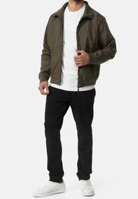 Veste bomber vert olive avec fermeture éclair à l'avant, poignets et col côtelés, associée à un pantalon noir et des baskets blanches, avec une coupe décontractée.