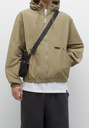 Personne portant une veste à capuche beige zippée, chemise blanche, pantalon noir et sac bandoulière noir avec sangle traversant la poitrine.