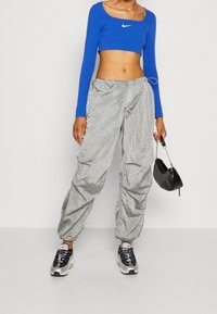 Blå långärmad kort topp med vitt logotyp, kombinerad med lösa, glänsande silvriga byxor och svarta sneakers, hållande en liten svart väska.