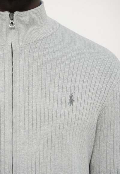 Szürke pamut cipzáras pulóver bordázott textúrával, magas gallérral és kis hímzett logóval a bal mellrészen. Cipzárzáródás elöl.