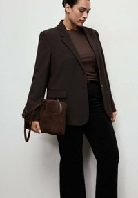 Blazer marrón combinado con una blusa marrón y pantalones negros. Lleva un bolso pequeño marrón con un acabado texturizado y detalles de cremallera.