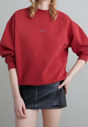 Osoba ubrana w czerwoną bluzę Holzweiller Oslo z podwiniętymi rękawami oraz krótką czarną skórzaną spódnicę, z ręką na biodrze.