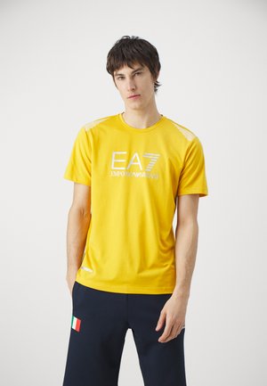 T-shirt in cotone giallo con colletto rotondo, stampa del logo EA7 Emporio Armani bianco e accenti a strisce sulle spalle. Tessuto morbido, maniche corte.
