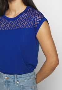 Top azul de manga corta con un panel de hombro texturizado y estampado; tejido suave; corte relajado; combinado con shorts de mezclilla azul claro.