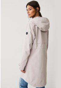 Chaqueta impermeable gris claro con capucha, que presenta un detalle de botón negro en la manga y una abertura en la espalda para mejorar la transpirabilidad.