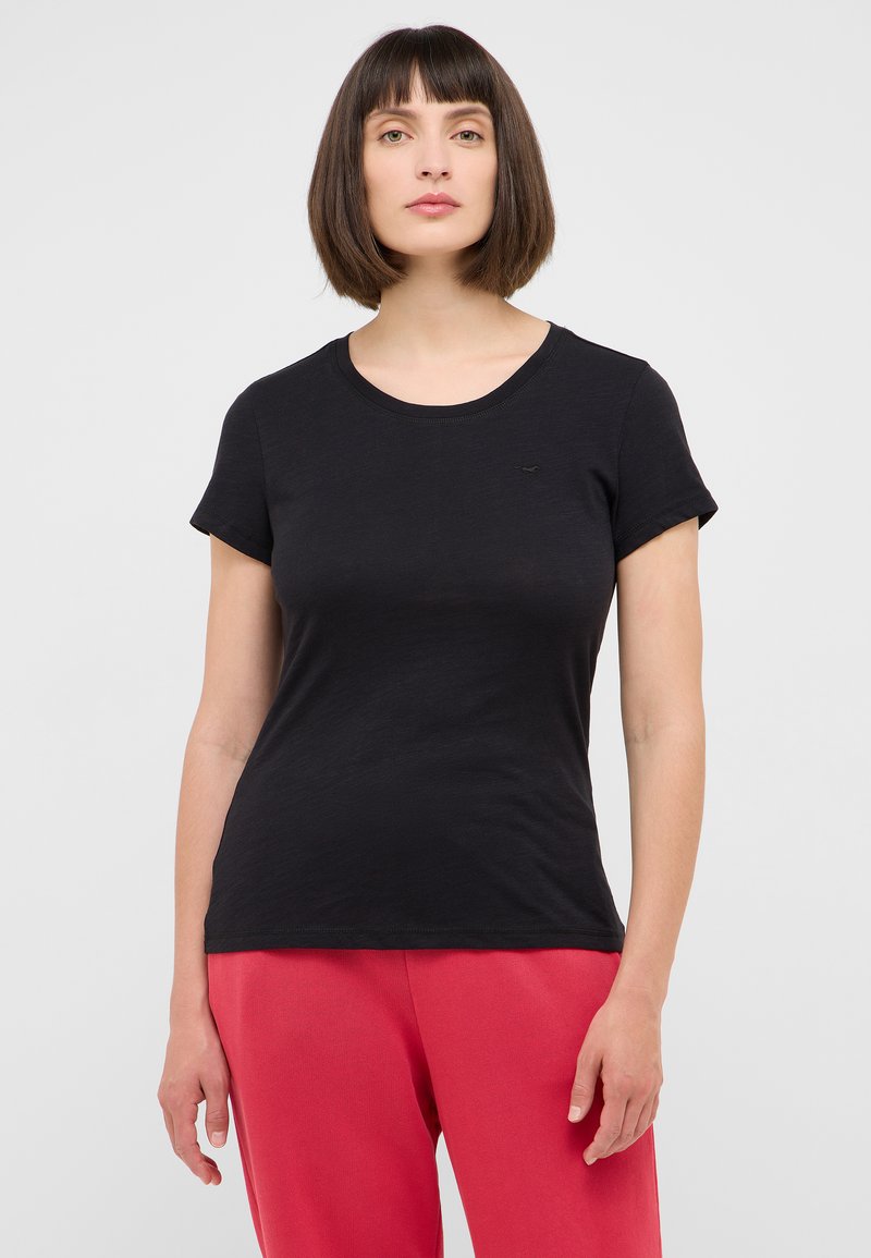 T-shirt noir à manches courtes, col rond, en tissu doux, design ajusté, détail logo subtil sur la poitrine, associé à un pantalon rose.