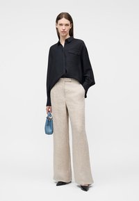 Γυναίκα που φοράει φαρδιά μαύρη μπλούζα, φαρδιά μπεζ παντελόνια, μαύρα παπούτσια, κρατώντας μια μικρή μπλε τσάντα, στέκεται μπροστά σε απλό φόντο.