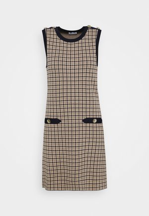Nainen pukeutuneena kamelintakkiin, jonka päällä on laivastonsininen ja beige hanhenjalkakuosinen mekko ja mustat loaferit, joissa on kultaiset ketjuyksityiskohdat.