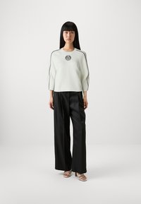 Maglione bianco a costine con accenti neri e logo, abbinato a pantaloni larghi neri e sandali con tacco beige. Il modello è in piedi contro uno sfondo neutro.