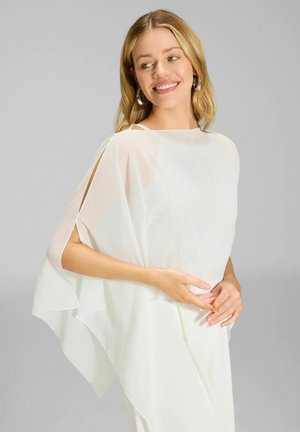 Blonde Frau trägt ein fließendes weißes Oberteil mit offenen Ärmeln, lächelt und schaut seitlich vor einem grauen Hintergrund.