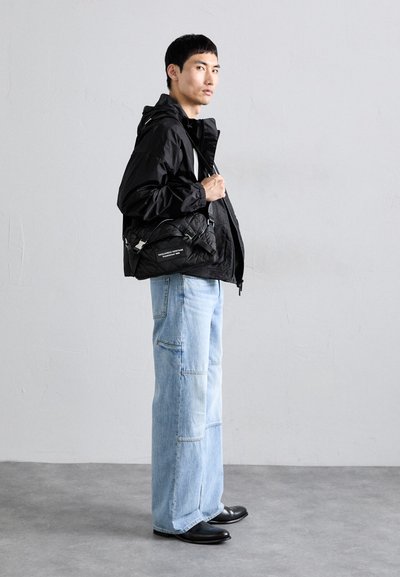 Schwarze Nylonjacke mit Reißverschluss und Kapuze, kombiniert mit hellblauen weit geschnittenen Jeans mit Patches. Schwarze Lederschuhe runden den Look ab.