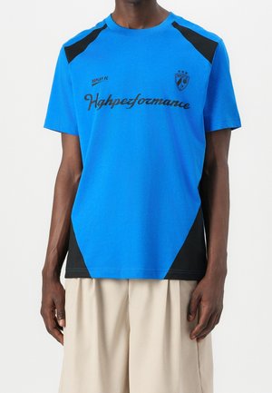 Osoba ubrana w niebieski t-shirt z krótkim rękawem z czarnymi akcentami, z nadrukiem "Highpezformance" i małym emblematem na piersi, w połączeniu z beżowymi spodniami.