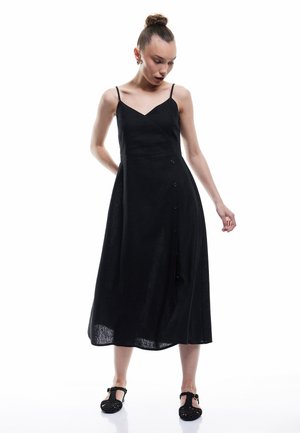 Czarna sukienka na ramiączkach w kształcie litery V, z dopasowaną górą i rozkloszowaną spódnicą, ozdobiona guzikami po boku. Teksturowana tkanina.