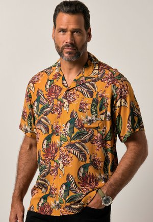 Mann mit Bart, der ein gelbes kurzärmeliges Hemd mit grünem, braunem und weißem tropischem Blumenmuster trägt, steht mit einer Hand in der Tasche.