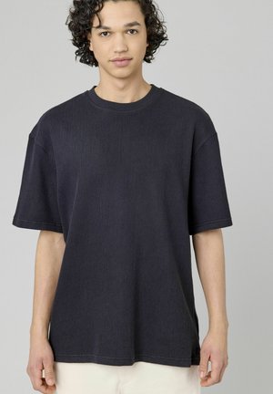 T-shirt à manches courtes, de couleur bleu marine foncé, avec une texture côtelée, un col rond et une coupe décontractée. Coutures apparentes et fentes sur les côtés.