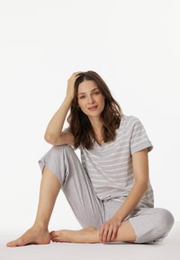 Siva prugasta majica s v-neck izrezom i svijetlosive capri hlače, izrađene od mekanog materijala. Model pozira ležerno s jednom rukom u kosi, bosa.