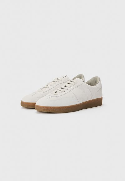 Weiße Wildledersneaker mit glatter Oberfläche, runder Zehenpartie, Schnürdesign und brauner Gummisohle. Dezente Logodetails an der Seite.