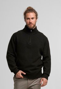 Zwarte sweater met ritssluiting en geribde kraag en manchetten, gemaakt van gestructureerd breimateriaal, gedragen met lichtgekleurde broeken.