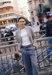 Sivi krojeni blazer preko belega topa, kombiniran z ohlapnimi sivimi kavbojkami. Mesto v ozadju z motorji in dekorativno arhitekturo.