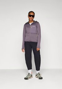 Ženska v vijoličnem zip-up jopiču, črnih kratkih hlačah, belih nogavicah, sivih športnih copatih in velikih črnih sončnih očalih, ki stoji ob enobarvni podlagi.