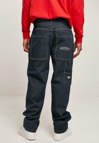 Pantaloni cargo in denim blu scuro con doppie tasche posteriori, ricamo del logo, cuciture a contrasto e vestibilità comoda. Abbinati a una camicia rossa a maniche lunghe.