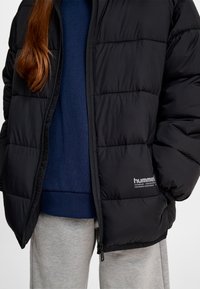 Person iført en sort quiltet jakke, marineblå sweater og lysegrå bukser, med hår delvist synligt over skulderen.