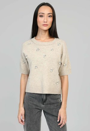 Pull en maille beige à manches courtes, avec des embellissements floraux et un col rond. Associé à un jean gris foncé.