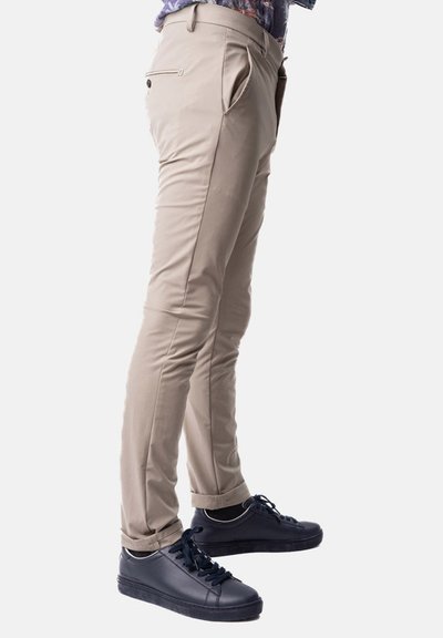 Pantaloni beige slim-fit realizzati in tessuto liscio, con tasche frontali e un dettaglio di tasca con zip. Indossati con sneaker blu scuro.