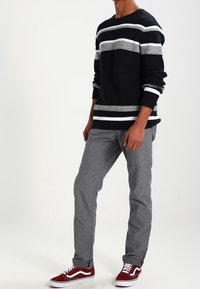 Maglione a righe navy con linee orizzontali grigie e bianche, abbinato a pantaloni grigio chiaro e sneakers rosse. Vista frontale, posa in piedi.