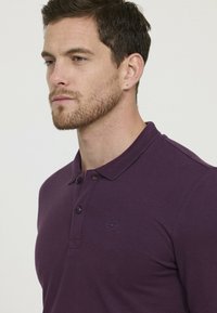 Polo shirt violet avec un col texturé, deux boutons et un logo circulaire sur la poitrine. Le tissu semble lisse et ajusté.