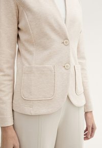 Blazer beige clair en tissu texturé, avec deux poches avant et deux boutons. Coupe slim avec un col à revers et un design ajusté.
