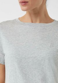 Jasnoszara koszulka z krótkim rękawem, wykonana z miękkiego materiału, z prostym, okrągłym dekoltem oraz małym logo w dolnym rogu.