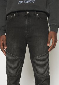 Black denim jeans featuring ribbed detailing on the thighs, standard pockets, and a button closure. Fabric has a slightly textured finish.
