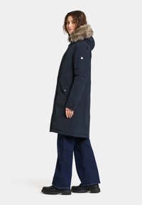 Laivaston pitkä takki, jossa on karvavuorella vuorattu hupullinen päällinen, vetoketju- ja napituskiinnitys, sivutaskut ja teksturoitu kangas; yhdistetty leveälahkeisiin farkkuihin ja mustiin saappaisiin.