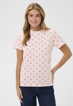 Jauna sieviete ar gariem blondiem matiem, valkā gaiši rozā T-kreklu ar maziem sarkaniem ķiršu rakstiem un tumši zilas džinsas, smaidot ar roku uz gurna.