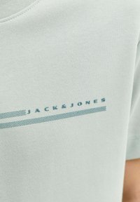 Tissu gris clair avec petit texte bleu « JACK & JONES » au-dessus de deux bandes pointillées parallèles au centre d’un vêtement.