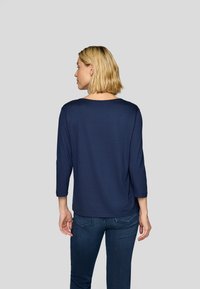 Marineblaue Bluse mit 3/4-Ärmeln und rundem Halsausschnitt. Weicher Stoff, lockere Passform, detaillierte Naht entlang der Rückennaht. Kombiniert mit dunklen Jeans.